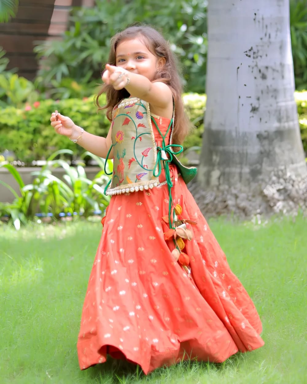 A girl in a coral lehenga with a painted sage bodice, mid-step on grass