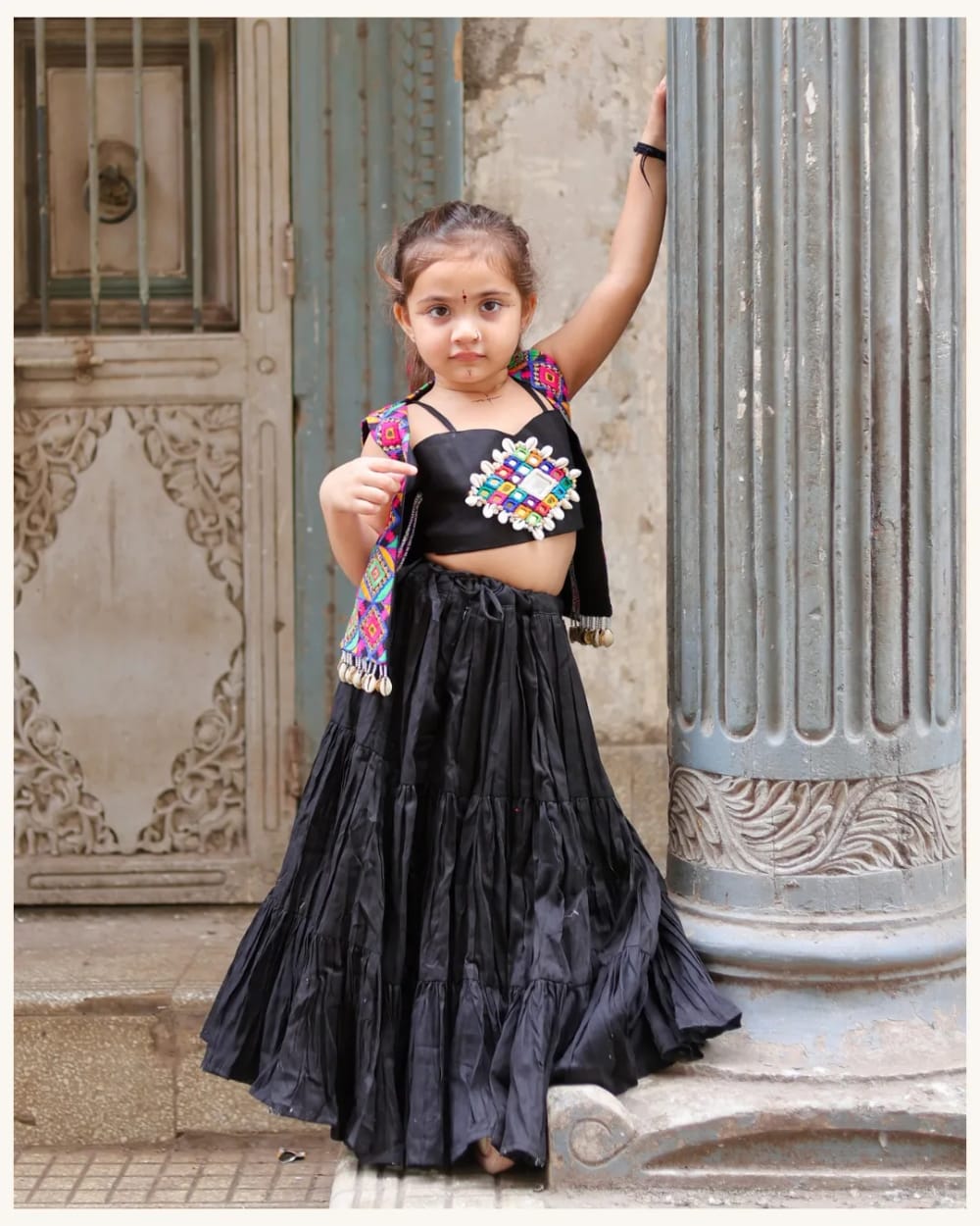 A girl in the black ghagra and Kutchi jacket, one arm raised against a carved wall